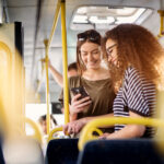 Two cheerful pretty young women are standing in a bus and looking at the phone and smiling while waiting for a bus to take them to their destination.